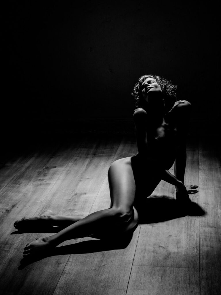 Boudoir shooting and nude photography, black and white, woman with curly hair and long legs sits sideways on the floor and looks relaxed upwards to the light :: photo copyright Karin Bergmann