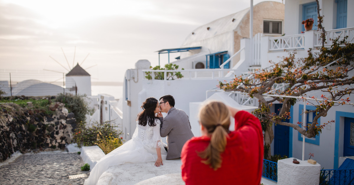 Wedding photo with bridal couple and wedding photographer in the picture; the couple kissing, picturesque surroundings with white houses and windmill, behind them the sea, making-of photo shoot on the island of Santorini, Greece:: photo copyright Karin Bergmann