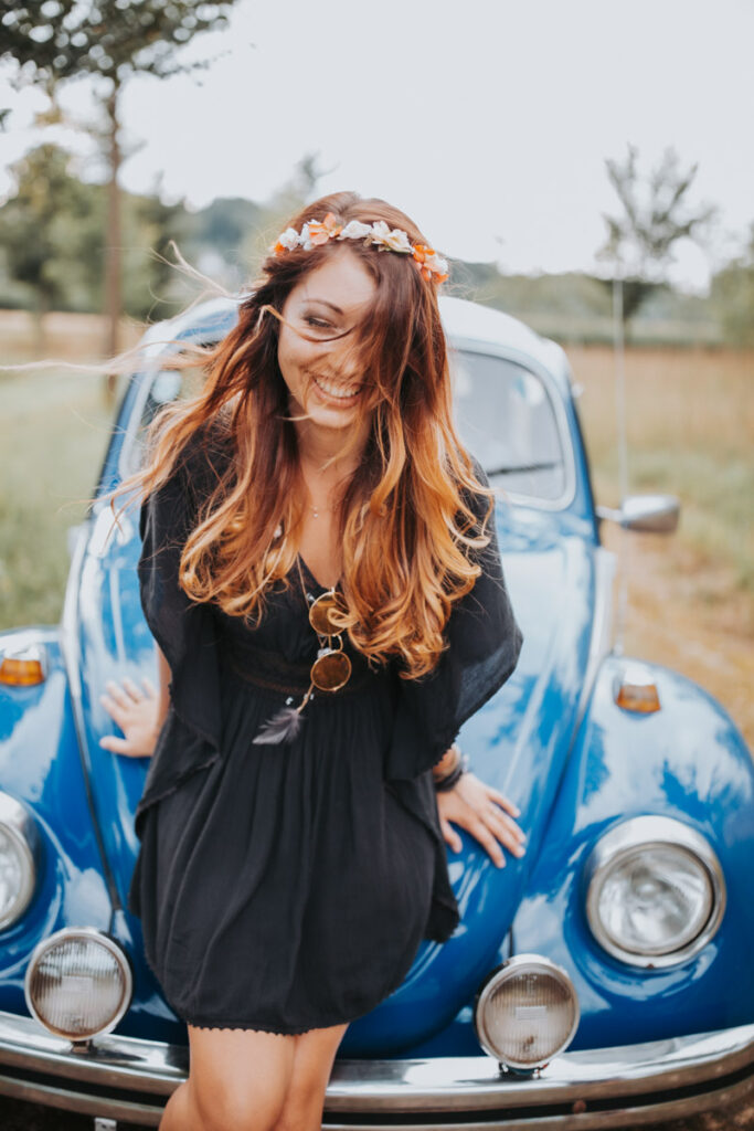 Lifestyle portrait, expressive photo shoot with young woman with long brown hair and a wreath of flowers on her head leaning against a blue Volkswagen Beetle on a meadow :: photo copyright Karin Bergmann