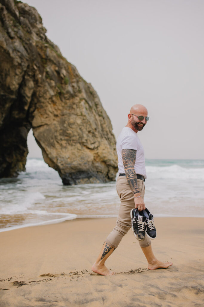 Lifestyle portrait, expressive, tattooed man walking on a beach in Portugal, behind him impressive cliff :: photo copyright Karin Bergmann