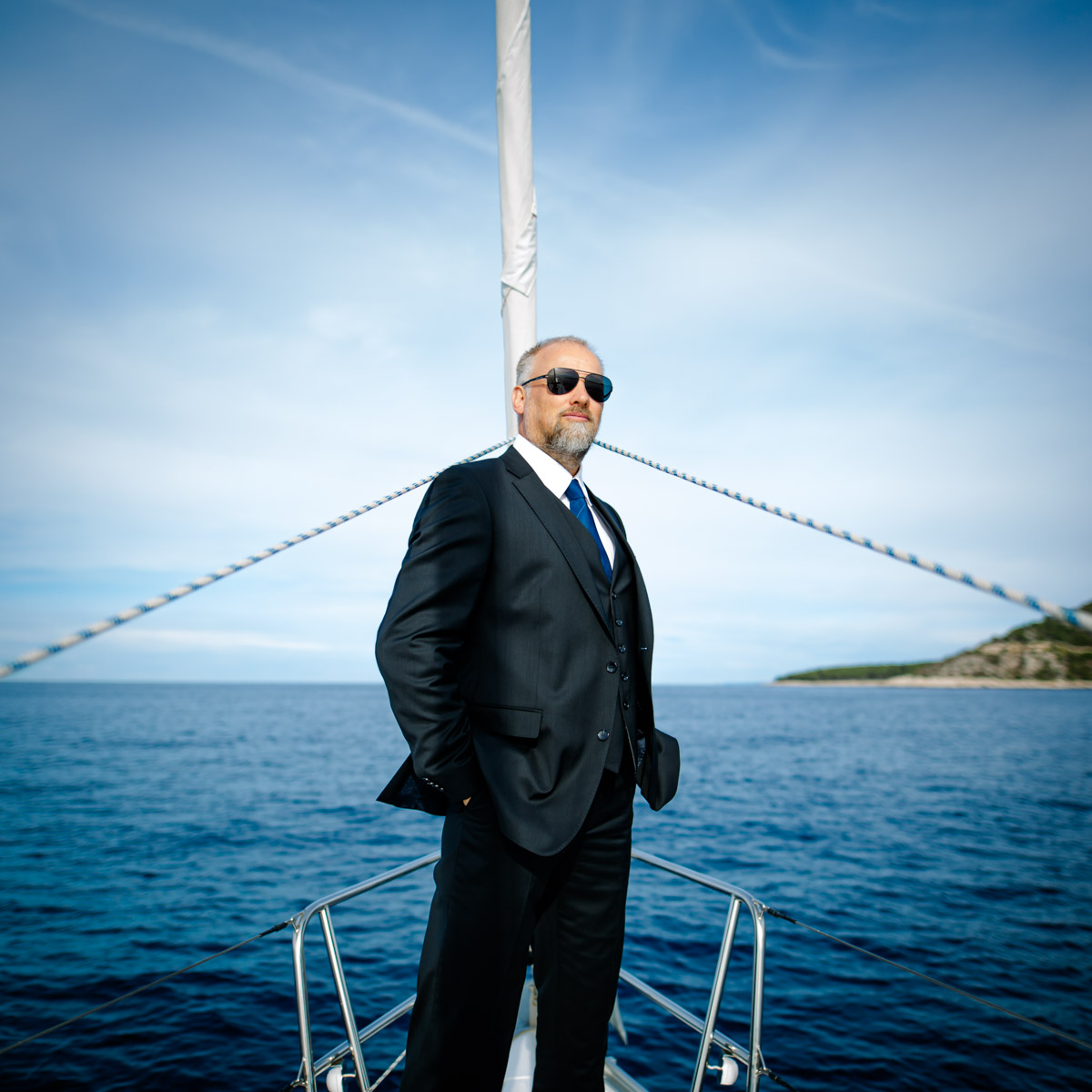 exklusives und ausdruckstarkes Lifestyle Portrait Fotoshooting, Mann im schwarzen Anzug mit Sonnenbrille steht am Bug einer Yacht, dahiner eine Insel und der Horizont des Meeres :: photo copyright Karin Bergmann